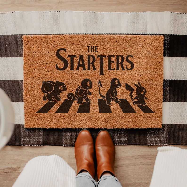 A top-down view of a rectangular natural coir doormat displaying four iconic retro anime characters walking across a street crossing with a person standing on it