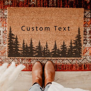 May include: A rectangular tan coir doormat featuring a black silhouette of a dense pine forest across the bottom half. The words Custom Text are printed in a simple black sans-serif font in the upper center area of the textured natural fiber mat.