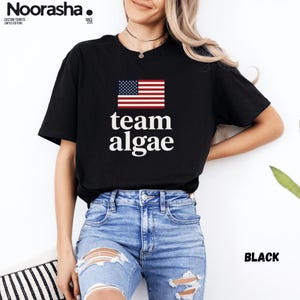 Peut inclure: Un t-shirt noir à manches courtes arborant un graphique du drapeau des États-Unis au-dessus du texte blanc "team algae". Le t-shirt est associé à un jean bleu clair présentant des déchirures usées sur les cuisses et les genoux.