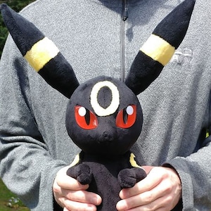 Könnte beinhalten: Schwarzes und gelbes Plüschtier eines Pokemon-Charakters mit roten Augen. Das Spielzeug wird von einer Person gehalten.