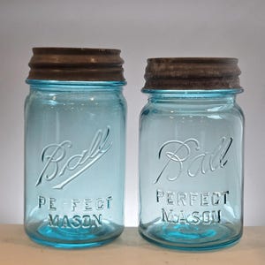 May include: Two vintage light blue glass jars with rusted metal lids. Each jar features the embossed script text Ball and the block text PERFECT MASON on the front. The glass has a distinct aqua tint and a cylindrical shape with a threaded neck.