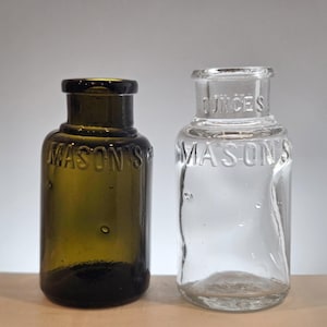 May include: Two small glass bottles with short necks and wide openings. One bottle is dark olive green and the other is clear glass. Both bottles feature the embossed text MASON'S on the front, and the clear bottle also has the word OUNCES embossed.