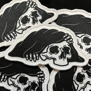 May include: A collection of black and white patches featuring a skull wearing a hood. The skull is holding its head with its hands. The patches have a white border.
