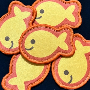 May include: A collection of yellow fish-shaped patches with orange trim and smiling faces. The patches are made of fabric and have an embroidered border. The background is a dark blue color.