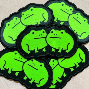 May include: A collection of embroidered patches featuring a bright green frog design on a black background. The frog patches have a simple, cartoonish style, with a minimalist design. The patches are likely for clothing or accessories.