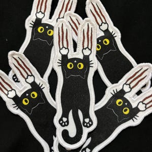 May include: A collection of black cat patches with yellow eyes and white outlines. The cats are depicted with their paws raised, claws extended, and red scratch marks. The patches are likely for clothing or accessories.