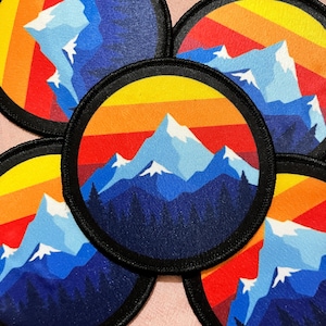 May include: Circular patches with a mountain landscape design. The patches feature a mountain range with snow-capped peaks, a forest, and a sunset gradient of yellow, orange, and red. Each patch has a black border.