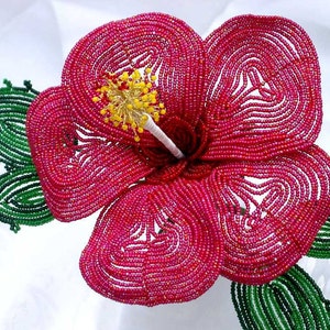 May include: A beaded hibiscus flower with pink petals and green leaves. The flower is made with small, round beads and has a yellow center.