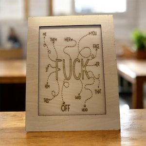 May include: Wooden framed art piece with the word "FUCK" in the center, surrounded by arrows and words like "ME", "YOU", and "OFF". The art is laser-etched into the wood, creating a rustic, humorous design.