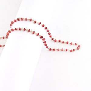 Puede incluir: Un delicado collar con pequeñas piedras preciosas rojas facetadas ensartadas en una cadena dorada. El collar se muestra sobre un fondo blanco, resaltando el color vibrante de las piedras preciosas y el brillo sutil de la cadena.