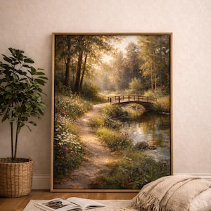 Op de afbeelding: Een ingelijst landschapsschilderij toont een serene bosscène met een houten brug over een beek. Het kunstwerk toont een pad dat de bossen in leidt, met zonlicht dat door de bomen filtert. Het schilderij wordt aan een muur getoond.