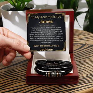 May include: A black leather bracelet with silver and gold accents, presented in a wooden box. The box contains a card with the text "To My Accomplished James" and a heartfelt message.