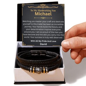 May include: Black leather bracelet with silver and gold accents, presented in a white gift box. The bracelet features a braided design and a magnetic clasp. A card inside the box reads "To My Hardworking Son Michael" with a heartfelt message.