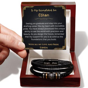 May include: A black braided leather bracelet set inside a wooden gift box. The bracelet has silver and gold-colored accents and the words "love you forever". A card inside the box reads "To My Accomplished Son Ethan".