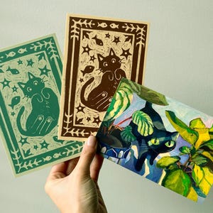May include: Three art prints featuring cat illustrations. One print is green with a cat and fish design, another is brown with a cat and star design, and the third is a colorful painting of a cat among plants.