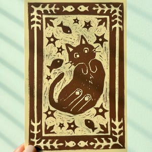 May include: A brown and beige art print featuring a cat lying on its back, surrounded by stars and fish. The print has a decorative border with leaves and fish. The cat has its paws up and is looking at the viewer.