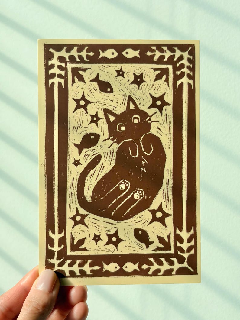 May include: A brown and beige art print featuring a cat lying on its back, surrounded by stars and fish. The print has a decorative border with leaves and fish. The cat has its paws up and is looking at the viewer.