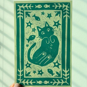 May include: A teal and cream-colored art print featuring a playful cat design. The cat is depicted with its paws up, surrounded by stars and fish. The print has a decorative border with floral and fish motifs.