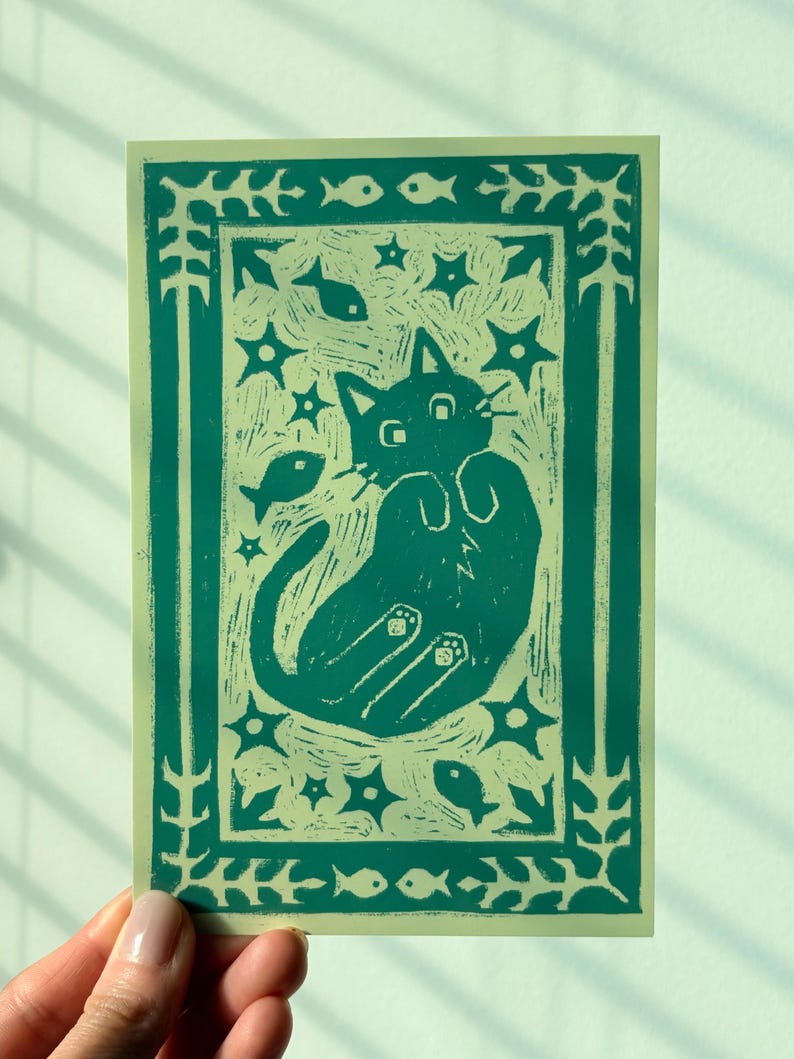 May include: A teal and cream-colored art print featuring a playful cat design. The cat is depicted with its paws up, surrounded by stars and fish. The print has a decorative border with floral and fish motifs.