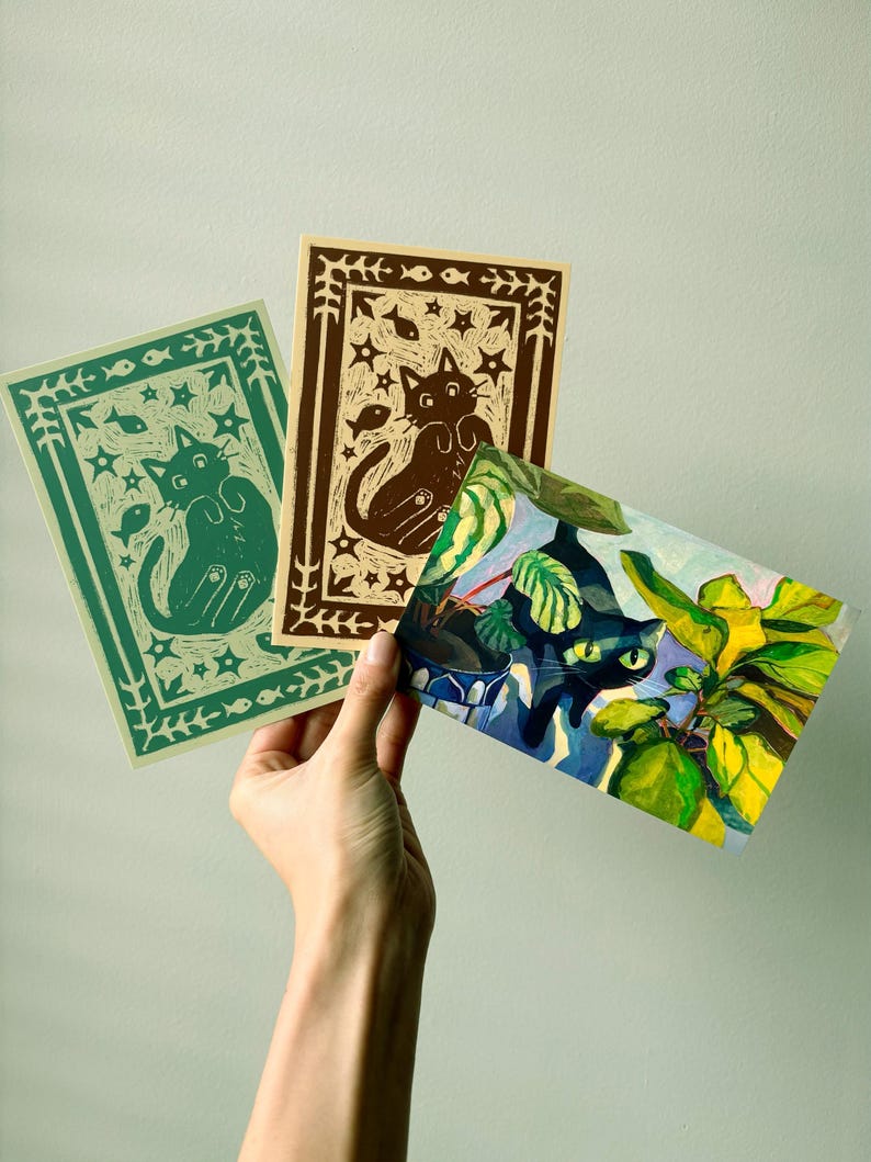 May include: Three art prints featuring cat illustrations. One print is green with a cat and fish design, another is brown with a cat and star design, and the third is a colorful painting of a cat among plants.