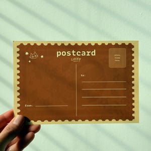 May include: A brown postcard with the word "postcard" in white, along with the word "LUCKY" below it. The postcard has a stamp-like border and includes spaces for "to:" and "from:" and a stamp area.