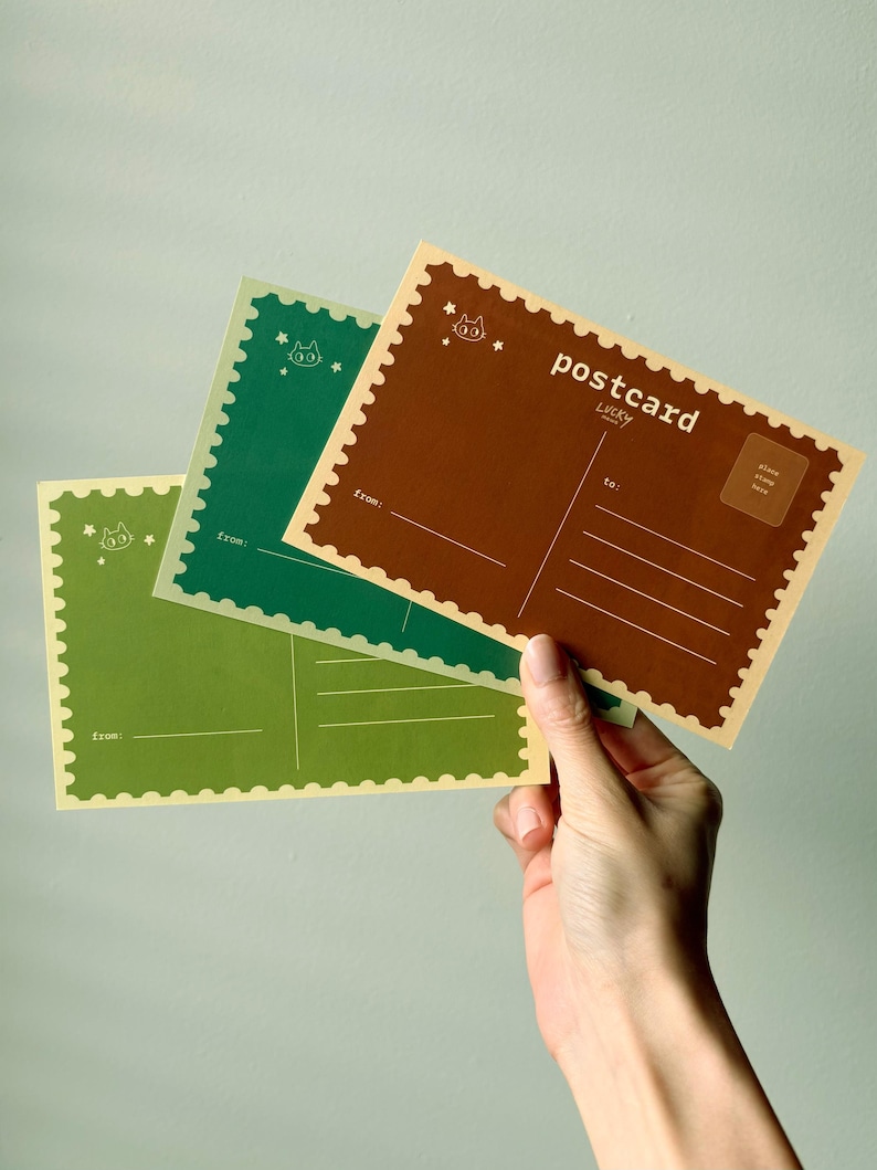 May include: Three postcards in shades of green and brown are held in a hand. The postcards have a scalloped edge and feature a cat illustration and the word "postcard". The back of the postcards have lines for writing and a space for a stamp.