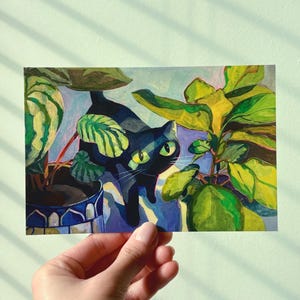 May include: A colorful illustration of a black cat peeking through lush green and yellow leaves. The cat has bright green eyes and is surrounded by potted plants in a blue and white pot. The artwork has a whimsical, artistic style.