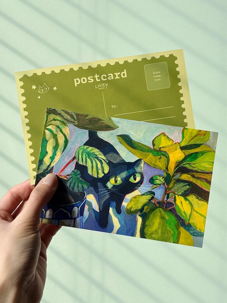 May include: A postcard with a painting of a black cat with green eyes amongst green and yellow plants. The postcard has the word "postcard" printed on it, along with the word "LUCKY". The background is a light green colour.