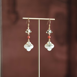 Puede incluir: Pendientes dorados con un colgante en forma de trébol. Cada pendiente presenta un trébol blanco, una cuenta roja y un acento floral con detalles blancos y verdes. Los pendientes se exhiben sobre un soporte dorado sobre un fondo marrón.