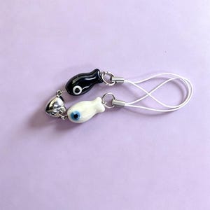 May include: A phone charm featuring two fish-shaped beads, one black and one white, each with a central eye design. A metallic silver heart-shaped bead is attached to the black fish. The charms are connected by silver jump rings to white cords.