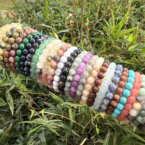 May include: A collection of beaded bracelets in various colors, including green, black, white, purple, brown, blue, and orange. The bracelets are made of gemstones and are displayed against a backdrop of green foliage.