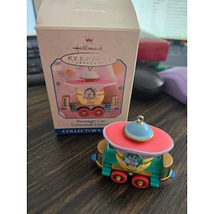 May include: A Hallmark Keepsake Ornament of a passenger car from the Cottontail Express. The ornament is a miniature train car with a red and blue top, green base, and gold accents. The box is visible in the background.