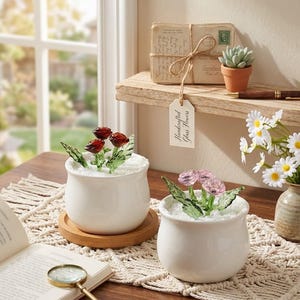 May include: Two white ceramic pots with handcrafted glass flowers. One pot has red roses, the other has pink flowers. The pots sit on a wooden table with a macrame runner, a book, and a magnifying glass. A tag reads "Handcrafted Glass Flowers."