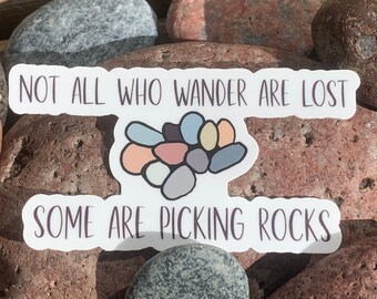 May include: White sticker with the text "Not all who wander are lost, some are picking rocks."  The sticker features a colorful illustration of a pile of rocks.