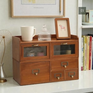 May include: A wooden desk organizer with multiple drawers and compartments. The organizer has a warm brown finish, glass-fronted drawers, and brass-toned hardware. A white mug, a glass jar, and a framed picture sit on top.