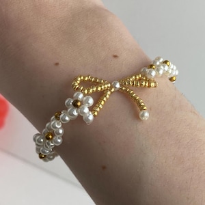 May include: A beaded bracelet featuring a gold bow and flower design. The bracelet is made of white and gold beads. The bow and flower are made of gold-colored metal. The bracelet is worn on a wrist.