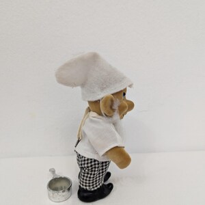 May include: A small, plush chef figurine wearing a white chef's hat and shirt, black and white checkered pants, and black shoes. A small metal cup sits beside the figurine. The figurine has a white beard and tan skin.