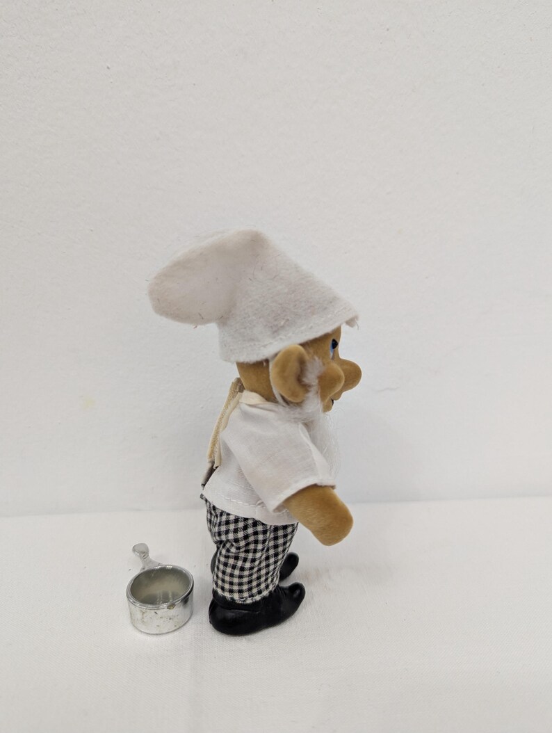 May include: A small, plush chef figurine wearing a white chef's hat and shirt, black and white checkered pants, and black shoes. A small metal cup sits beside the figurine. The figurine has a white beard and tan skin.