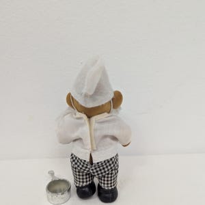 May include: A small chef figurine, viewed from the back, wearing a white chef's hat and shirt, and black and white checkered pants. A small silver metal pot sits on the surface next to the figurine.