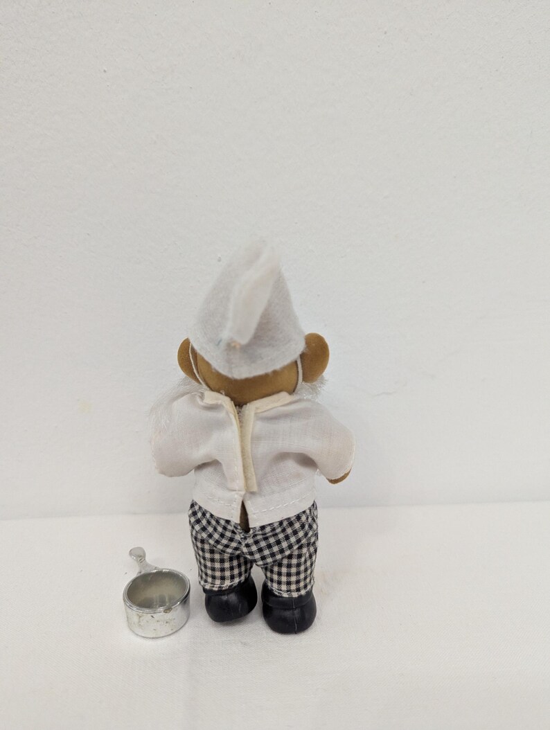 May include: A small chef figurine, viewed from the back, wearing a white chef's hat and shirt, and black and white checkered pants. A small silver metal pot sits on the surface next to the figurine.