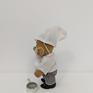 May include: A small, brown teddy bear figurine dressed as a chef, wearing a white hat, shirt, and a checkered pants. The bear has a white beard and is holding a small metal pot. The figurine is standing on a white surface.