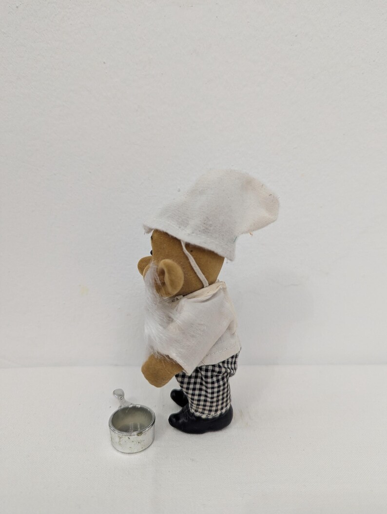 May include: A small, brown teddy bear figurine dressed as a chef, wearing a white hat, shirt, and a checkered pants. The bear has a white beard and is holding a small metal pot. The figurine is standing on a white surface.