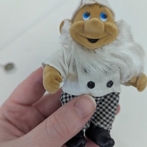 May include: A small, plush gnome figurine wearing a white chef's hat and jacket with black buttons. The gnome has a long white beard, a smiling face, and checkered pants. Black shoes complete the outfit.