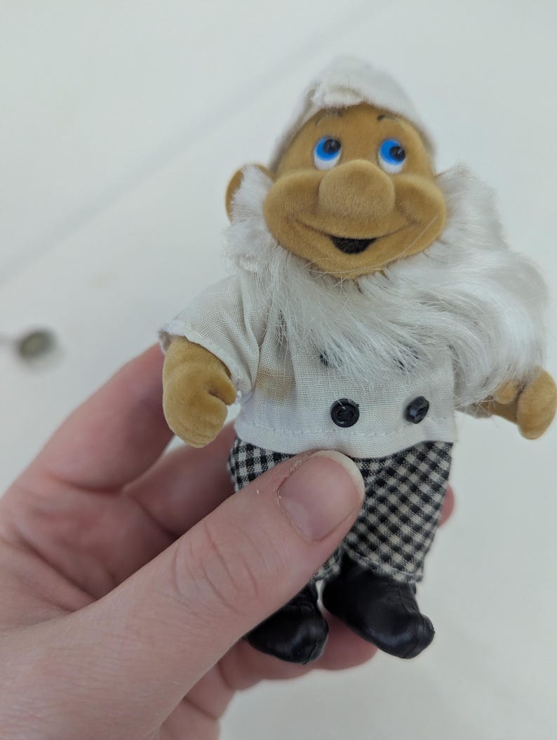 May include: A small, plush gnome figurine wearing a white chef's hat and jacket with black buttons. The gnome has a long white beard, a smiling face, and checkered pants. Black shoes complete the outfit.