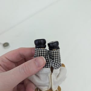 May include: A small, brown teddy bear doll wearing a white chef's coat and black and white checkered pants. The doll has black shoes. The doll is being held upside down.