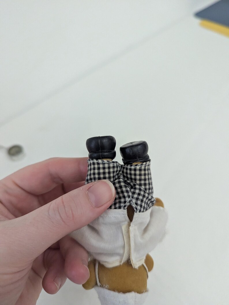 May include: A small, brown teddy bear doll wearing a white chef's coat and black and white checkered pants. The doll has black shoes. The doll is being held upside down.