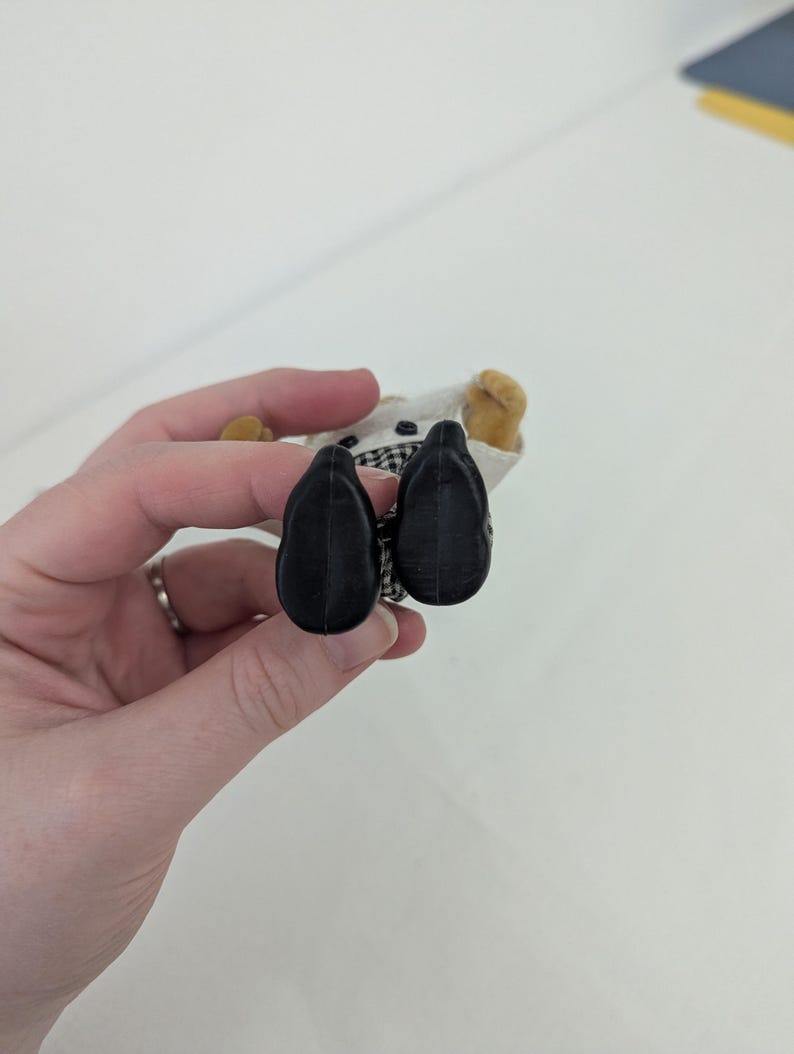 May include: A miniature doll wearing a chef's uniform, featuring a white jacket with a black and white checkered collar. The doll has black shoes and tan hands. The doll is held against a white background.