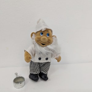 May include: A plush chef gnome doll wearing a white hat, shirt, and checkered pants. The gnome has a long white beard, blue eyes, and black shoes. A small silver metal measuring cup is next to the gnome.