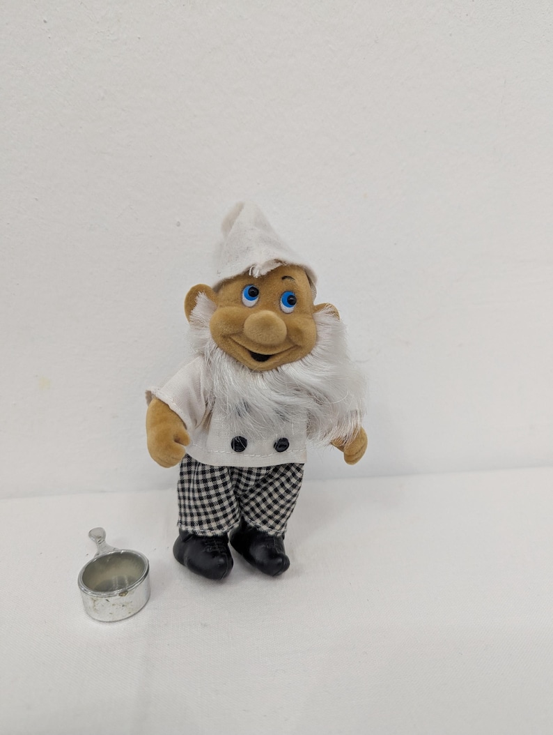 May include: A plush chef gnome doll wearing a white hat, shirt, and checkered pants. The gnome has a long white beard, blue eyes, and black shoes. A small silver metal measuring cup is next to the gnome.