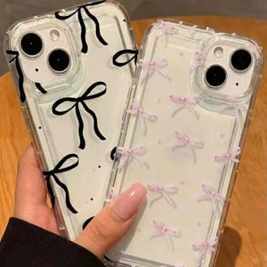 Peut inclure: Deux coques de téléphone transparentes. L'une présente un motif de nœuds noirs, l'autre de petits nœuds roses. Les coques sont tenues devant une surface marron clair.